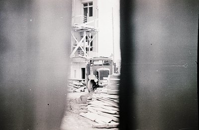 Construction scene: a truck stands amidst lumber and scaffolding before a multi-story building. Likely a mid-century commerci...