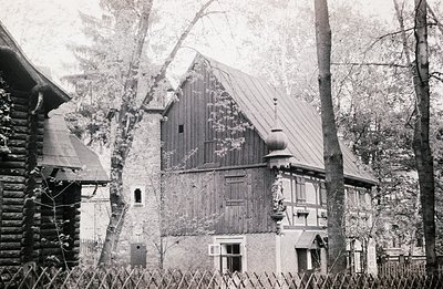 A timber-framed house with a steep, slate roof and decorative gables sits nestled amongst trees. A log cabin-style structure ...