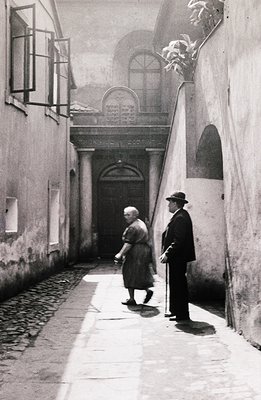 A monochrome street scene depicting an elderly couple walking toward a grand, arched doorway set within a stone facade. Paved...