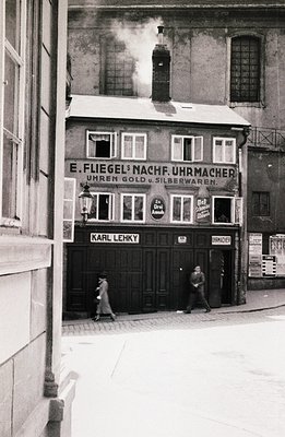B&W street scene captures a storefront "E. Fliegel's Nachf. Uhrmacher" (watchmaker) with ornate signage & a vintage lamp. Two...