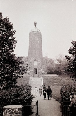 Monochrome photo depicts a stone tower with a conical roof and sculpted relief, ascending stone steps. Three figures, two adu...