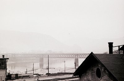 Overcast skies and fog obscure a railway bridge spanning a wide river, likely industrial in nature. A weathered building with...