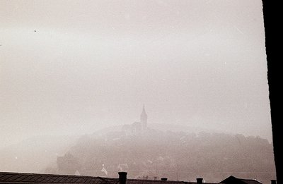 Ethereal cityscape view through a window frame. A hill crowned with a church spire dominates the scene, shrouded in mist or f...