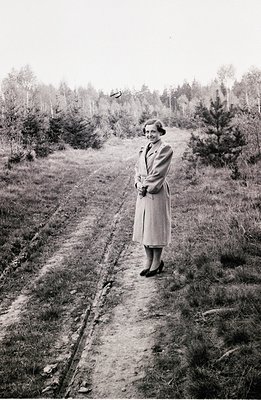 A woman in a tailored coat and heels stands on a dirt track, framed by pine trees in a forested area. Likely 1950s style; the...