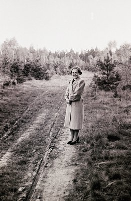 Elegant woman in a long coat poses on a dirt path leading into a pine forest. Mid-century style clothing and hairstyle sugges...