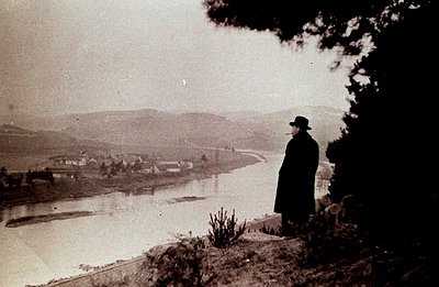 A solitary man in a dark coat and hat stands overlooking a winding river valley, likely a scenic vista. The photograph's grai...