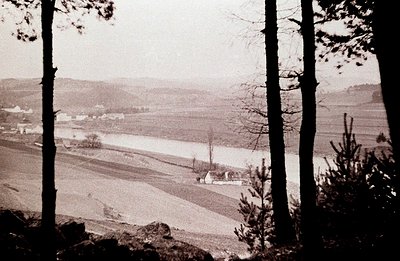 Undulating landscape with a wide, shallow river and small settlement visible through framing trees. Agricultural fields exten...