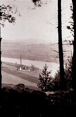 A monochrome view shows a modest building beside a wide, placid body of water, likely a lake or inlet. Gentle, terraced slope...