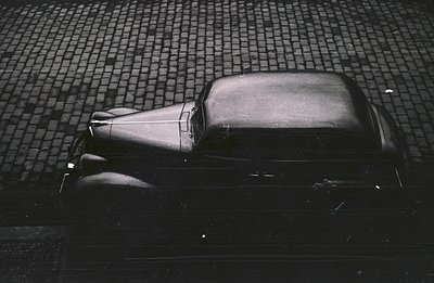 Classic car, likely a 1930s model, photographed from above on a cobbled street. The vehicle's dark color and angular design a...