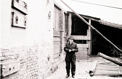 A young man in a dark suit and hat stands outside a weathered brick and timber building. The structure shows signs of age and...