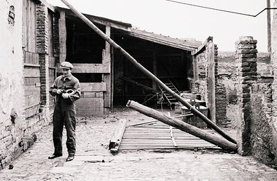Man in work overalls and cap stands in a brick courtyard, examining a small object in his hands. Weathered brick walls and a ...