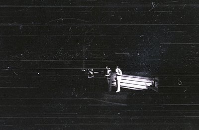 Three figures stand near a rectangular structure, possibly a platform or stage, partially obscured in darkness. The scene app...