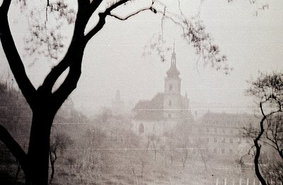 A view of a historic townscape, likely European, shrouded in mist. A church spire dominates the scene, visible above rooftops...