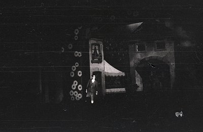 A monochrome image shows a person standing near a building featuring a detailed balcony with ornate railings and a partially ...