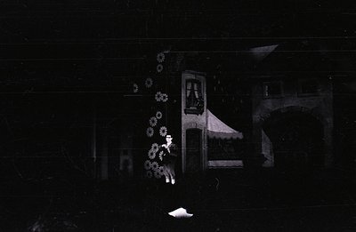 A young woman stands before a building facade, likely a theatrical set. The structure includes a balcony, arched entryway, an...