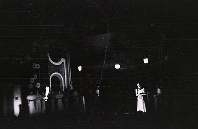 Eerie scene of a theatrical performance, possibly outdoors. A figure in white stands center, surrounded by draped fabric & st...