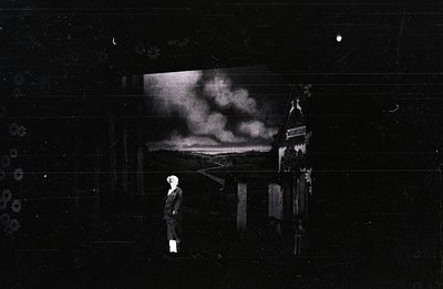 A solitary figure in period attire stands near a damaged building with a panoramic vista behind. Appears to document post-war...