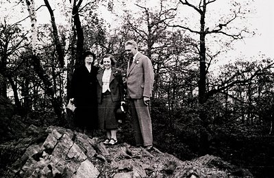 Three figures pose on a rocky outcrop amidst dense trees. A woman in a dark suit and hat stands to the left; a younger woman ...