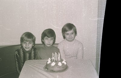 Three boys, likely siblings, sit behind a cake with lit candles. The image shows a mid-century style interior. Clothing sugge...