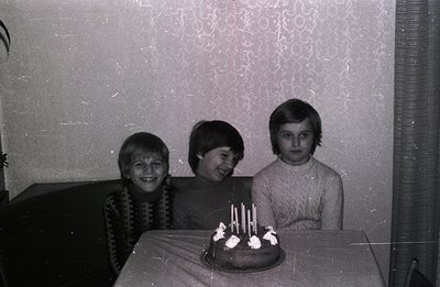 Three young boys, likely siblings, pose with a chocolate cake adorned with lit candles in a domestic setting. Visible details...