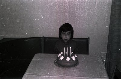 A young boy sits at a table covered with a tablecloth, focused on a birthday cake with seven lit candles. The room features t...