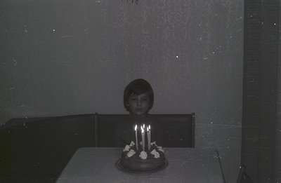 A young person sits behind a table covered with a tablecloth, facing a cake with seven candles. The room's wallpaper features...