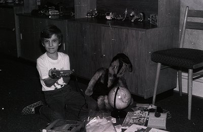 A young boy sits amidst toys and books, clutching a toy rifle. He's positioned in front of a dark wood cabinet displaying min...