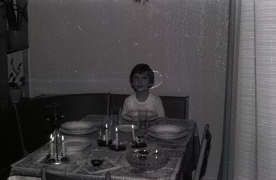 A young boy sits at a formally set table with multiple courses and lit candles. Textured wallpaper and dark furnishings sugge...