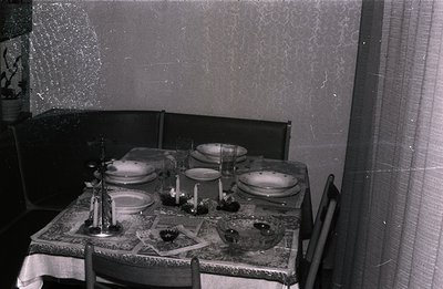 A formally set dining table occupies the scene, covered with a patterned tablecloth. Four place settings feature plates, glas...