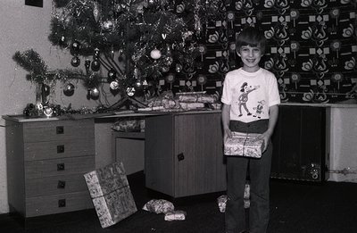A young boy, likely in the 1970s, stands proudly holding wrapped gifts in a festive setting. A fully decorated Christmas tree...