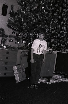 A young boy stands near a densely decorated Christmas tree, surrounded by wrapped presents. He wears a "Jetsons" themed t-shi...