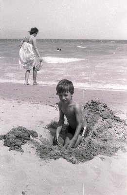 A young boy sits within a sand pit on a beach, looking toward the camera. A woman in a patterned dress stands further away, n...