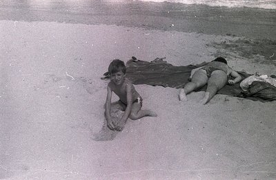Two boys in swim trunks relax on a sandy beach, one playing with sand, the other resting on a blanket. Grainy black and white...