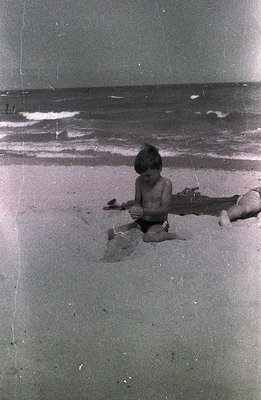 A young boy, lightly tanned and shirtless, kneels on a sandy beach, using a plastic shovel to build something. A log lies par...