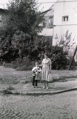 A young boy in shorts and a patterned shirt stands beside a woman with short hair, likely his mother. They pose on a cobbled ...