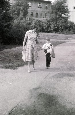 A woman and young boy walk hand-in-hand along a paved path. She wears a floral print dress; he carries a satchel. A large, mu...