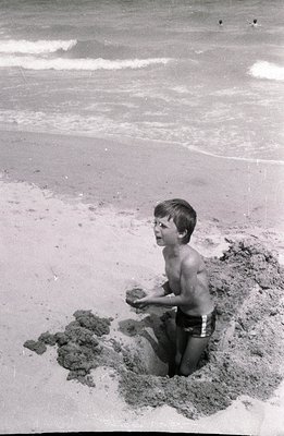 A young boy, possibly 7-10 years old, sits within a shallow pit he's dug on a sandy beach. He wears patterned swim trunks, ho...