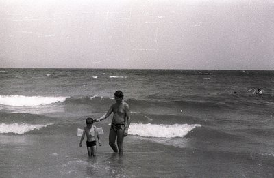 A man and young boy wade through shallow water along a sandy beach. The man holds the boy's hand. Waves gently break in the b...