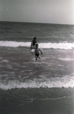 A boy wades into breaking waves, holding a large, inflatable beach toy resembling a swan. The image shows a mid-century beach...