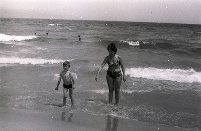 A mother and young son stand in shallow water along a sandy beach, waves gently lapping around them. The boy wears swim trunk...