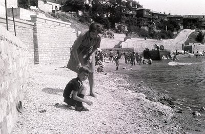 A young boy kneels on a pebbled beach, inspecting the ground, while a woman in a patterned dress watches nearby. A group of b...