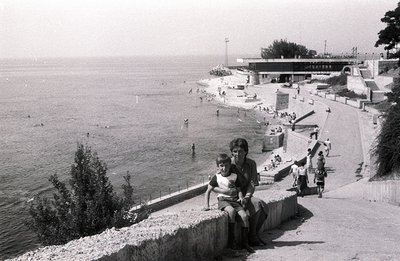 A family scene by the seaside: a mother and son sit on a stone retaining wall overlooking a sandy beach and the Black Sea. Th...