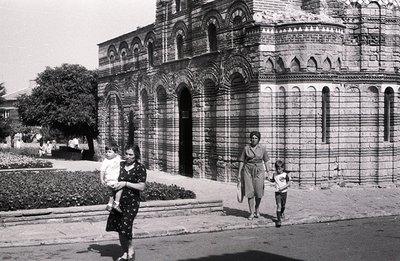 Striking b&w image: A family—mother carrying a child, another child running—pass before an ornate stone building with layered...