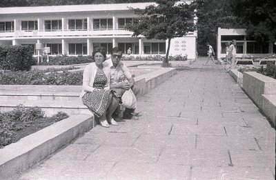 Black and white photo depicts a couple seated on a low wall bordering a formal garden path. Behind them, a modernist seaside ...