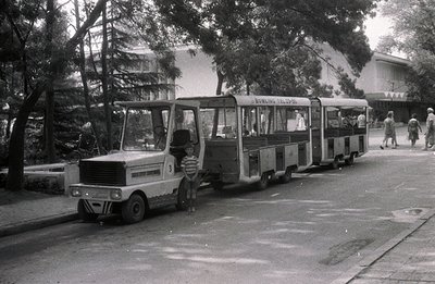 A vintage, open-air trolley or miniature train car stands on a paved road, a young boy stands near the front. The vehicle is ...