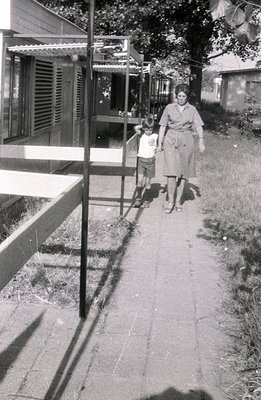 Black and white photo depicting a woman in a mid-length dress and a young boy walking along a brick-paved path. A building wi...