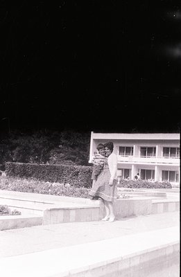 A woman carries a child outdoors in front of a modernist building, possibly a hotel or apartment complex. Features include a ...