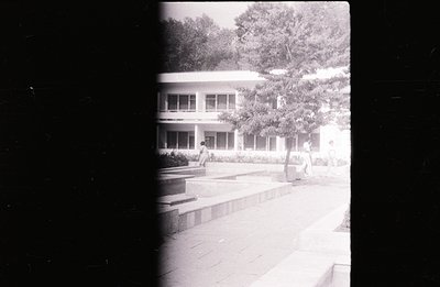 A monochrome photo showing a modernist building with large windows, viewed from a paved courtyard. Two figures are present, o...
