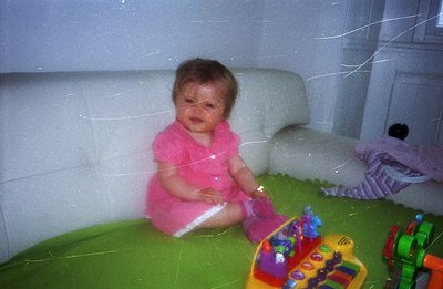 Infant sits on a green play mat, wearing a pink dress, surrounded by colorful toys. Appears to be a candid, indoor shot, like...