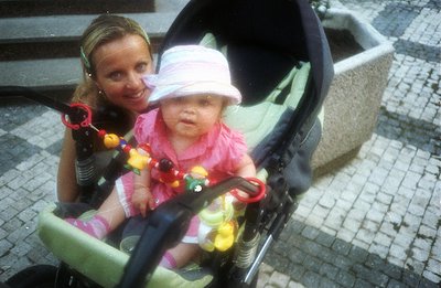 A baby in a stroller, wearing a pink hat and dress, gazes forward, held by a woman smiling in the background. A toy hangs fro...
